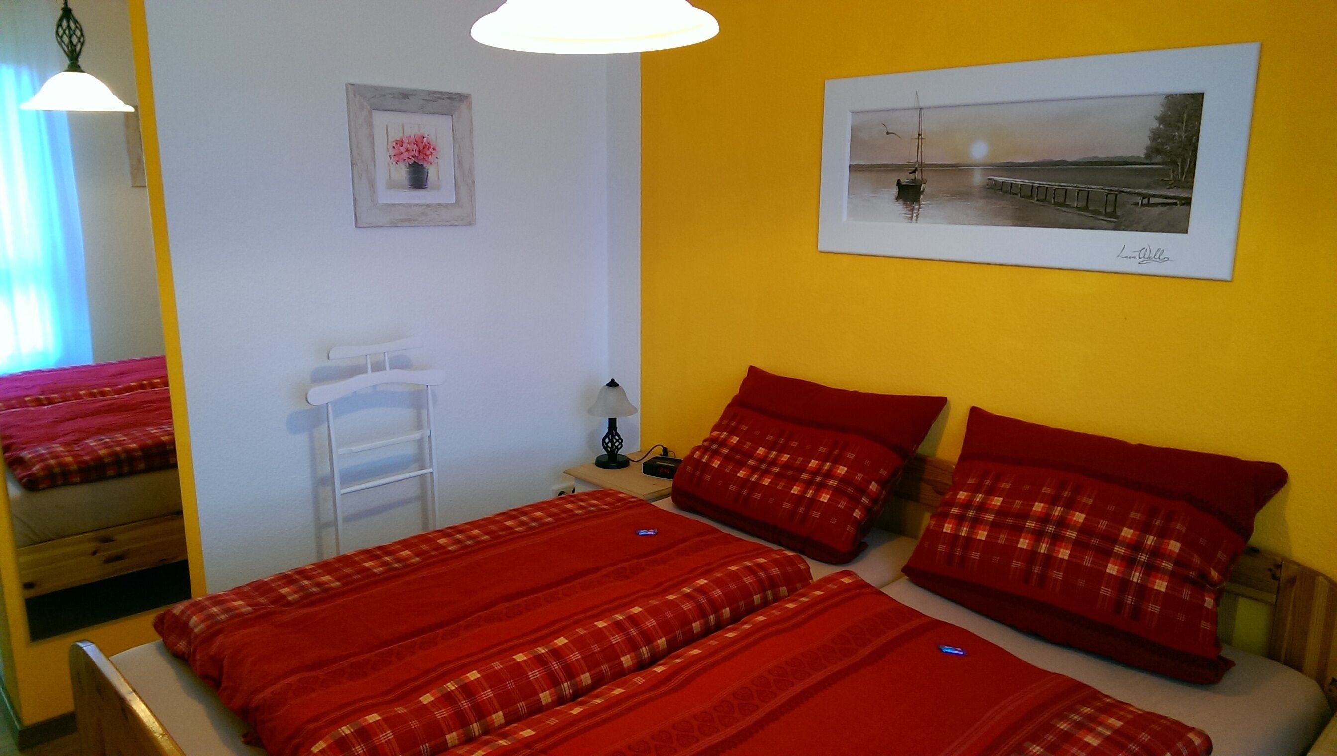 Ein Zimmer mit zwei Betten, jeweils mit roter Bettwäsche und einem Holzkopfende. An der Wand hängt ein gerahmtes Bild, ein weißer Heizkörper und ein kleiner Tisch mit einer Lampe.