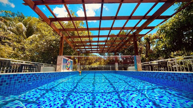 Una piscina cristallina blu con una pergola in legno sopra, circondata da vegetazione lussureggiante.