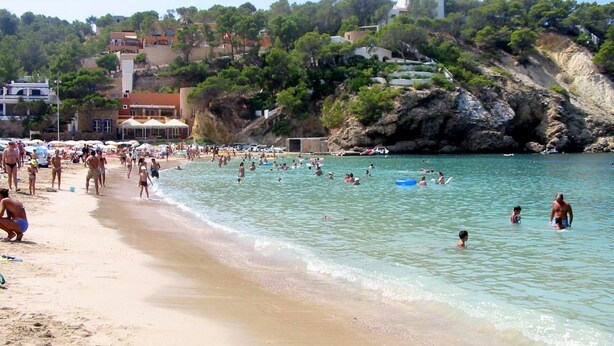 Una playa concurrida donde gente nada y toma el sol, con una orilla arenosa y edificios al fondo.