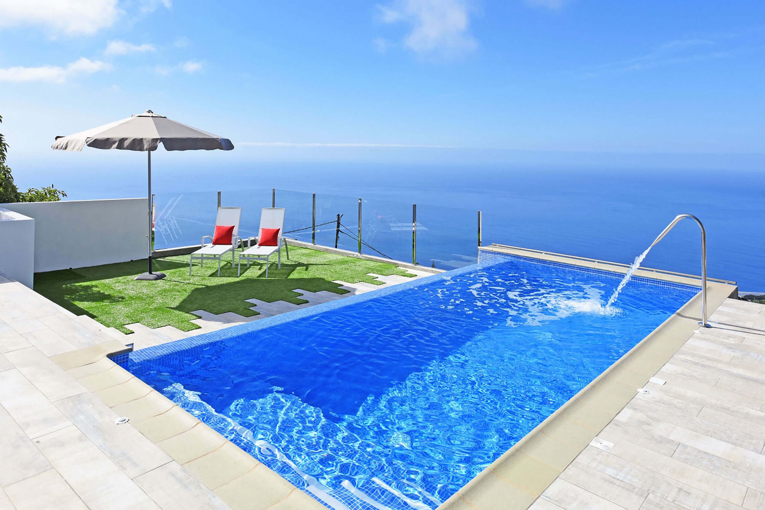 A swimming pool with a view of the ocean, a lounge area with chairs and an umbrella, and a small grassy patch.