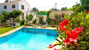 A swimming pool with clear blue water, surrounded by a garden with various plants and flowers. There are two lounge chairs and a small table with a potted plant. - CLASSIC ANDALUSIAN VILLA, SURROUNDED BY ORANGE TREES, FOUNTAINS. PRIVATE POOL. (Órgiva)