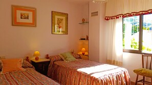 A room with two beds, a window with curtains, and a shelf with decorative items. - CLASSIC ANDALUSIAN VILLA, SURROUNDED BY ORANGE TREES, FOUNTAINS. PRIVATE POOL. (Órgiva)