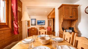 A wooden dining room with a table set for four, including plates, glasses, and cutlery. There are two chairs, a wooden cabinet, and a clock on the wall.