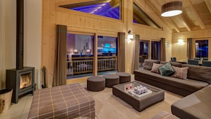 A cozy wooden interior with a fireplace, comfortable seating, and a large window overlooking a balcony.