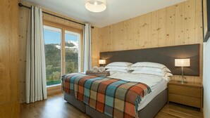 A bedroom with a wooden headboard, a large window with curtains, a bed with a plaid blanket, two bedside lamps, and a nightstand with a lamp.