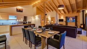 A spacious living area with a wooden ceiling, a sofa, a dining table set for four, and a kitchen in the background.