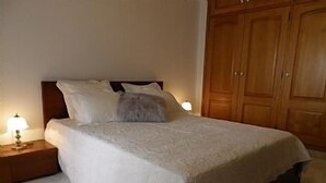 Una cama bien hecha con cubrecama y almohadas blancas, una mesita de noche de madera con lámpara, y un armario de madera en una habitación de hotel.
