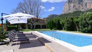 A swimming pool with lounge chairs and umbrellas, surrounded by greenery and a mountain backdrop.