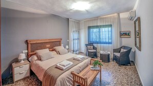 A hotel room with a large bed, a bedside table with a lamp, a chair, a small table with a plant, and a window with curtains.