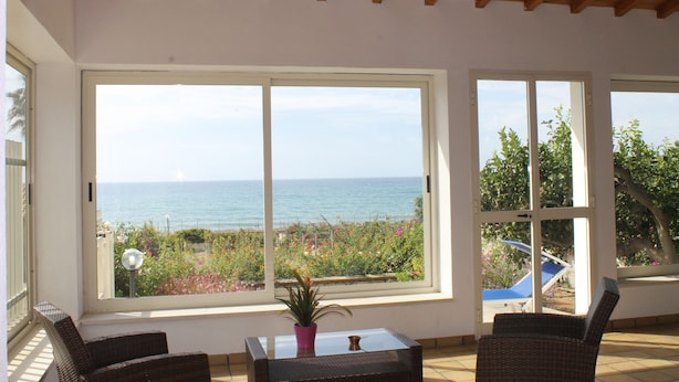 Una habitación con amplios ventanales con vista al mar, una planta en maceta sobre una mesa y mobiliario de mimbre.
