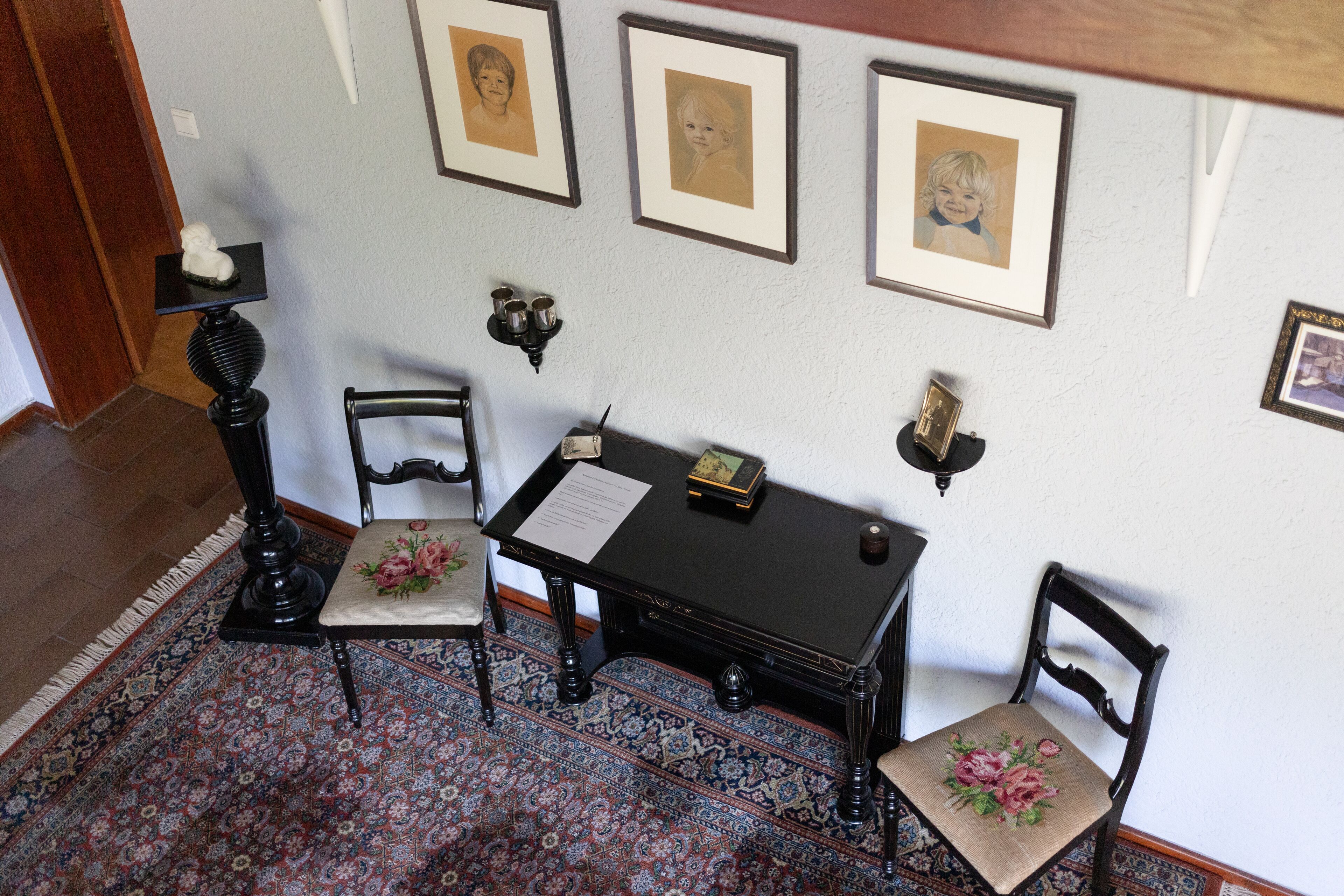 A room with a patterned rug, a desk with a lamp, chairs, and framed pictures on the wall.