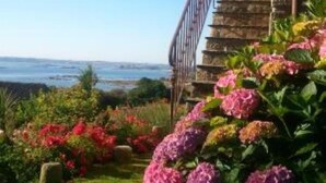 Ein Küstengarten mit einer Steintreppe, die zu einem Gebäude führt, umgeben von leuchtenden Blumen und freiem Blick auf das Meer.