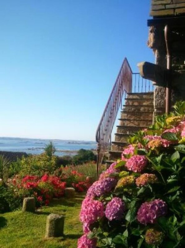 Un jardín costero con una escalera de piedra que conduce a un edificio, rodeado de flores coloridas y una vista despejada del océano.