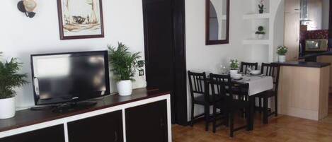 Ein Wohnzimmer mit Fernseher, Esstisch, Stühlen und Topfpflanzen.