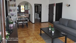 A living room with a grey sofa, a coffee table with potted plants, a dining area with a table and chairs, and a kitchen with a window and bookshelves.