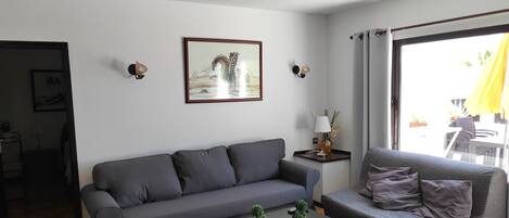 A modern living room with a grey sofa, a coffee table with two potted plants, a framed picture on the wall, and a large window with curtains.