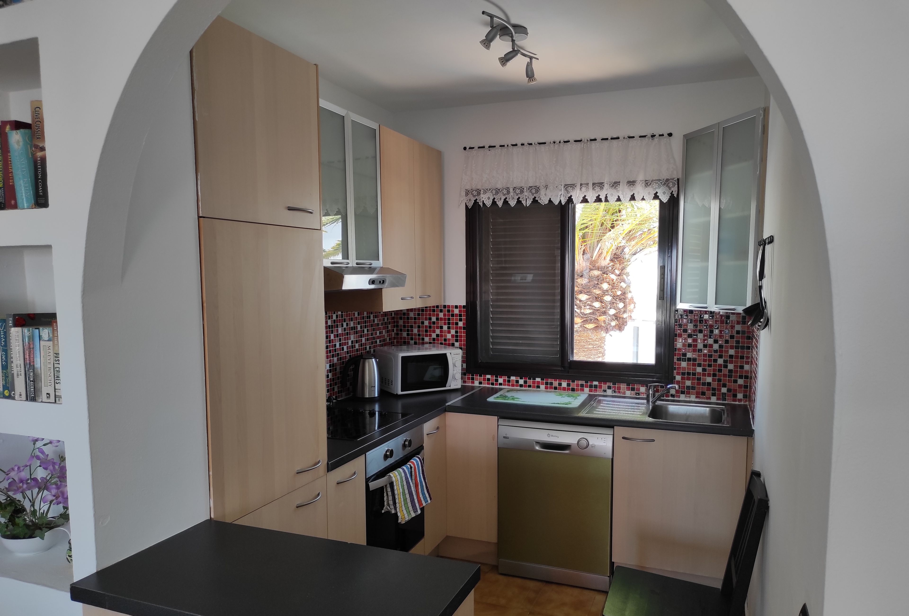 A compact kitchen with a microwave, stove, and sink. There is a window with blinds and a small plant on the left.
