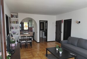 A living room with a grey sofa, a coffee table with potted plants, a dining area with a table and chairs, and a kitchen with a window and bookshelves. - Beautiful modern 2 bed apartment with a stunning sea view and close to the beach (Puerto del Carmen)