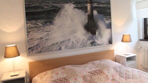 Ett sovrum med en stor tavla med havsmotiv, en sänggavel i trä, ett blommigt sängöverkast och två sänglampor.