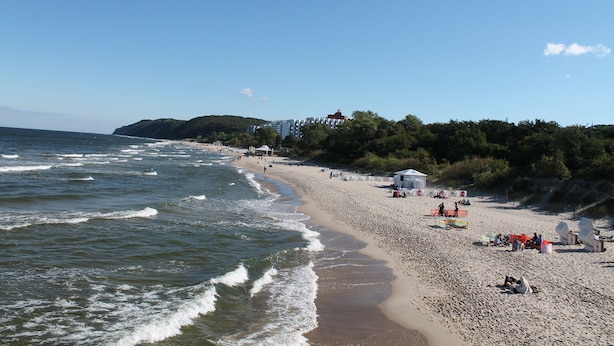 En strand med vågor, människor och byggnader i bakgrunden.