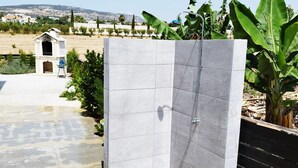 A modern outdoor shower area with a stone-tiled floor, a white tiled shower enclosure, and a view of a rural landscape in the background. - 5 Bed, 5 Bath Villa with Private Pool in Affluent Residence of Sea Caves (Paphos)