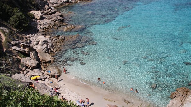 A rocky beach with clear turquoise water, people swimming and sunbathing.
