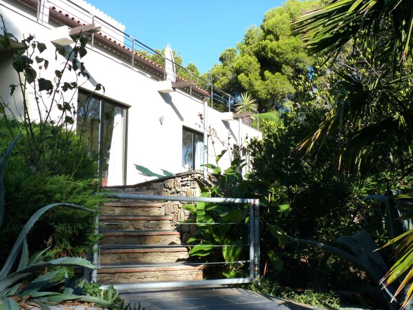 A white building with a stone staircase leading to a door, surrounded by lush greenery and a metal gate. - Luxurious house with huge private pool ( Begur)-  Free Wifi (Begur)