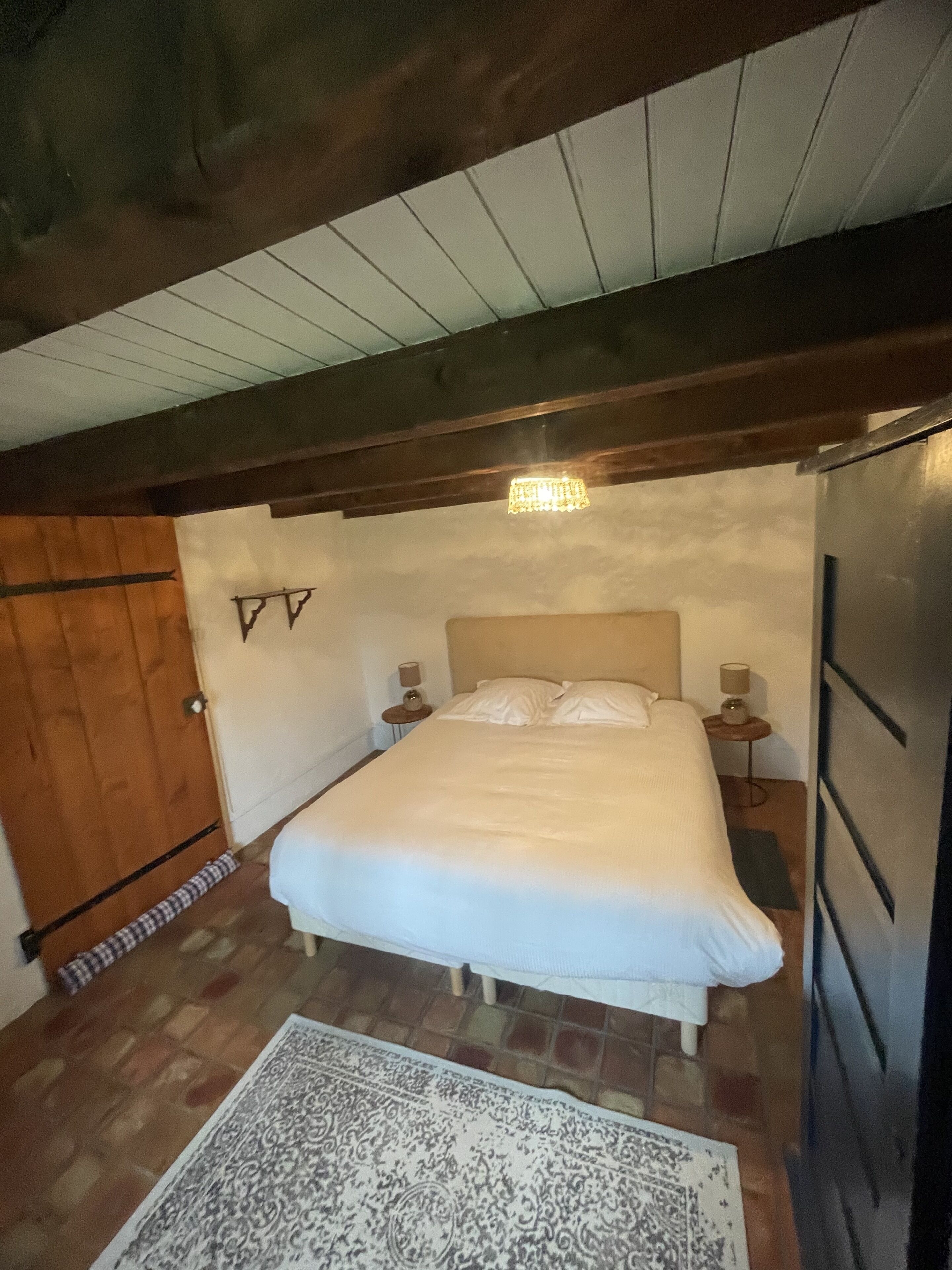 A bedroom with a large bed, wooden ceiling beams, a chandelier, and a refrigerator.