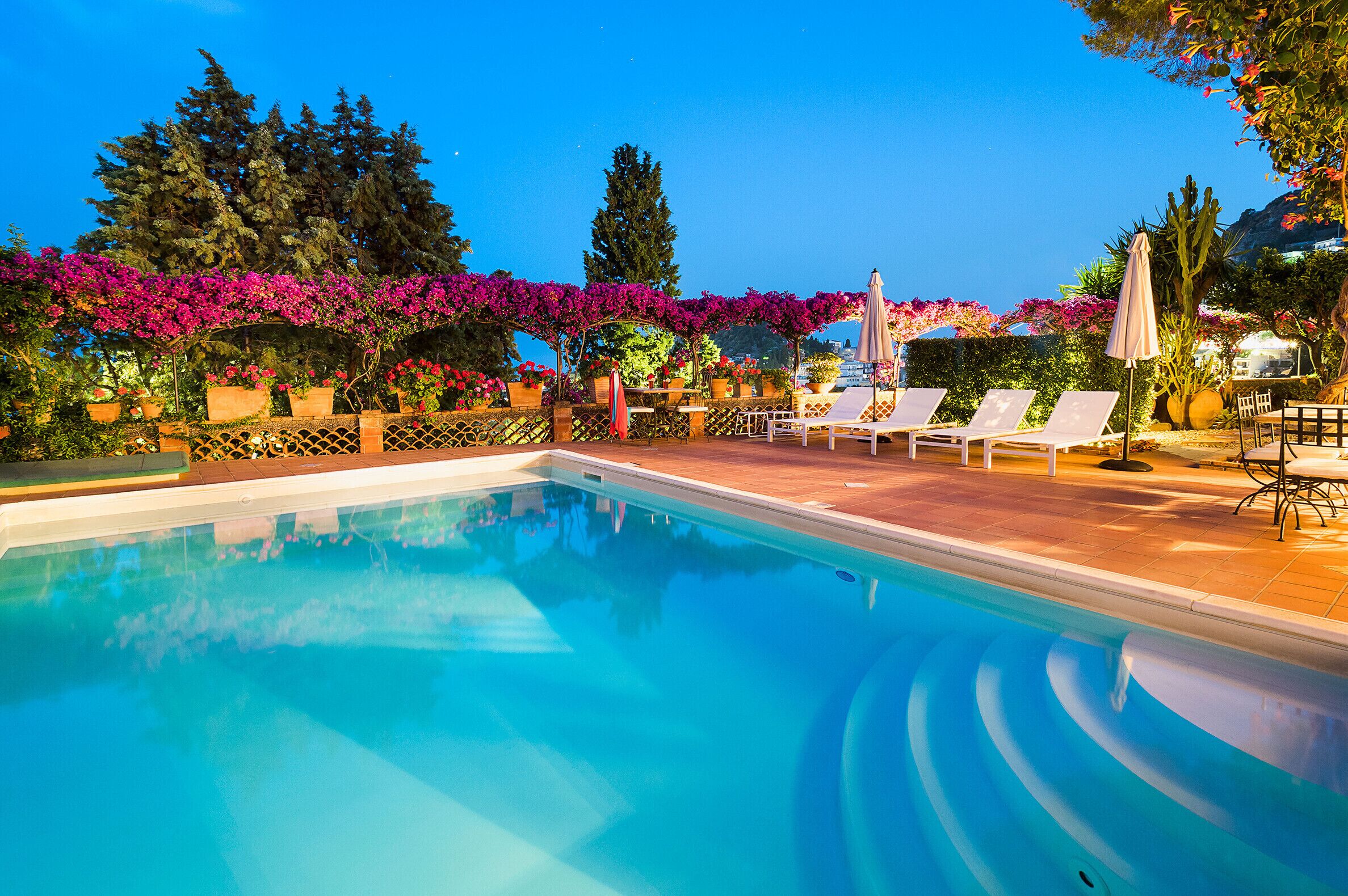 Piscina con sillones de descanso y sombrillas, rodeada de exuberante follaje y plantas con flores.