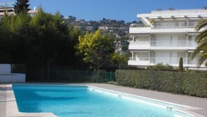 Uma piscina com deck ladrilhado, cercada por um hedge e um edifício com varandas.