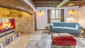 Ein gemütliches Wohnzimmer mit Kamin aus Stein, einem blauen Sofa, einem roten Hocker und einem Bücherregal.