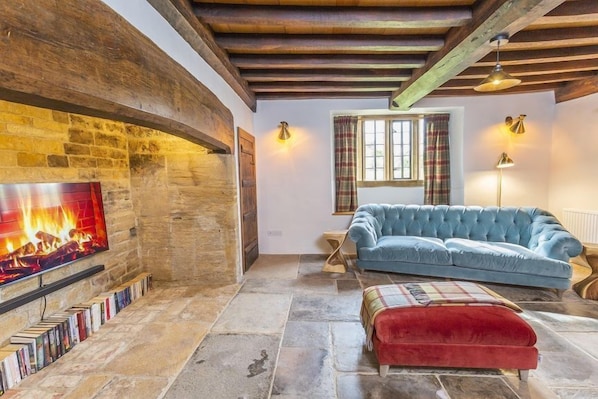 A cozy living room with a stone fireplace, a blue sofa, a red ottoman, and a bookshelf. - Historic Farmhouse with style (Martock)