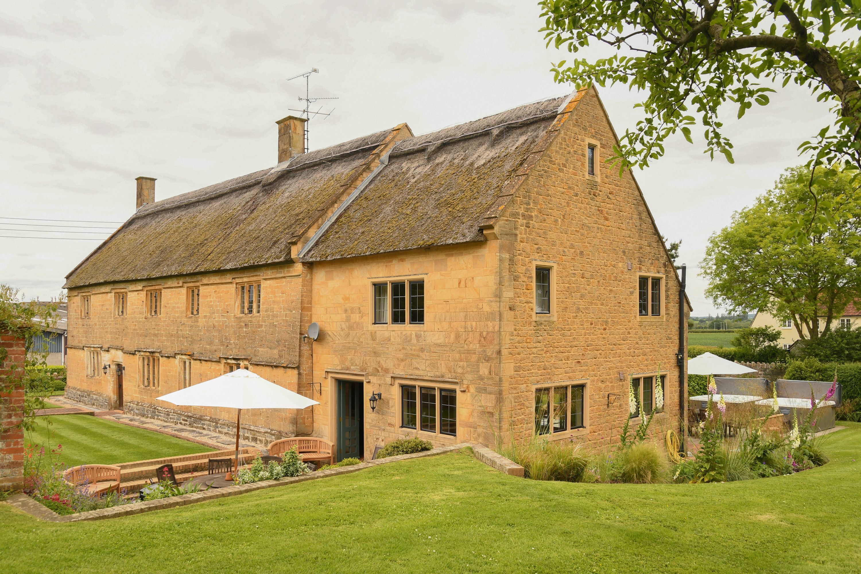 A stone building with a thatched roof, a garden with a bench, and a white umbrella.