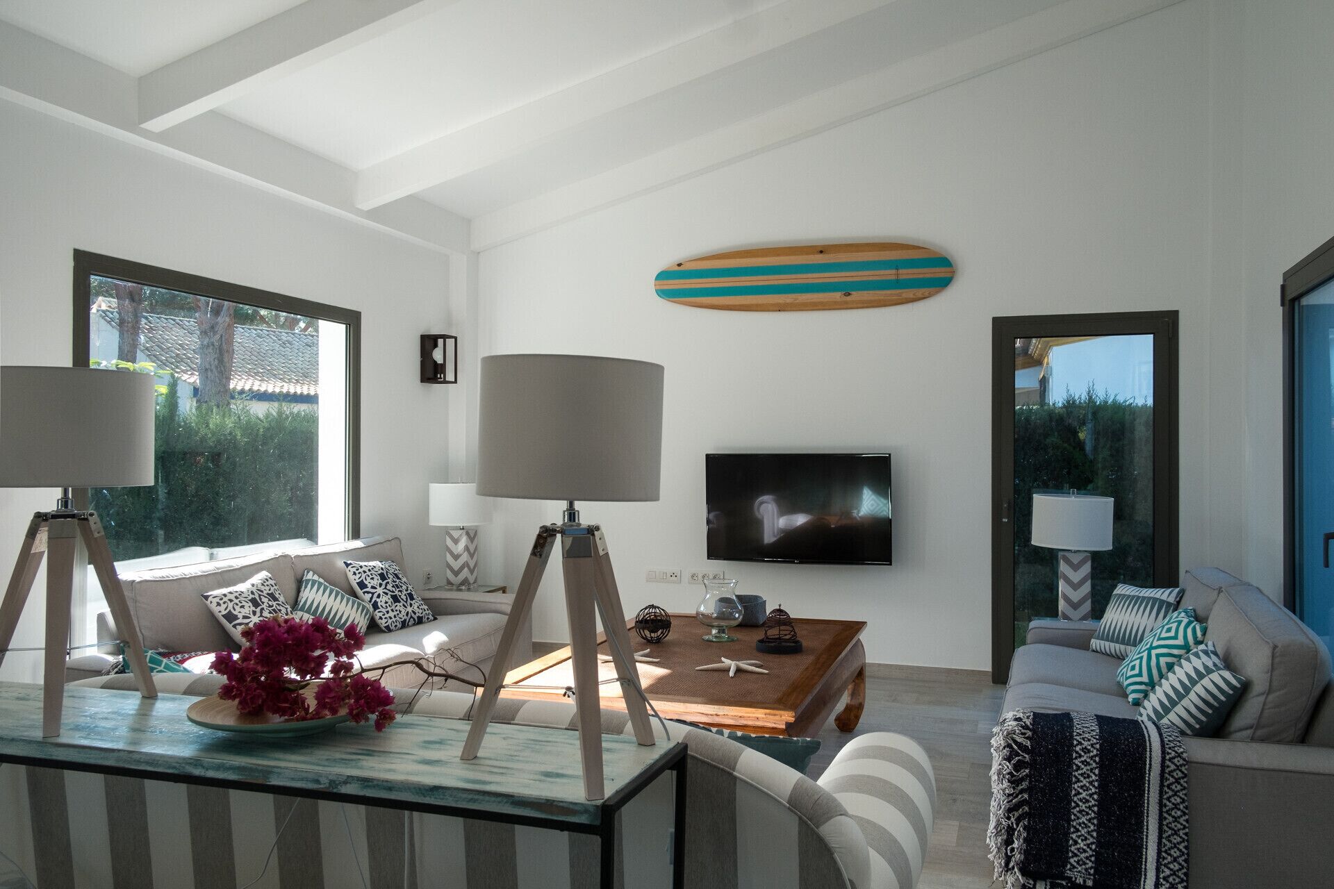 Un salon moderne avec un canapé, une table basse et un téléviseur. On y trouve deux lampes, un bol décoratif rempli de fleurs et une planche de surf accrochée au mur.
