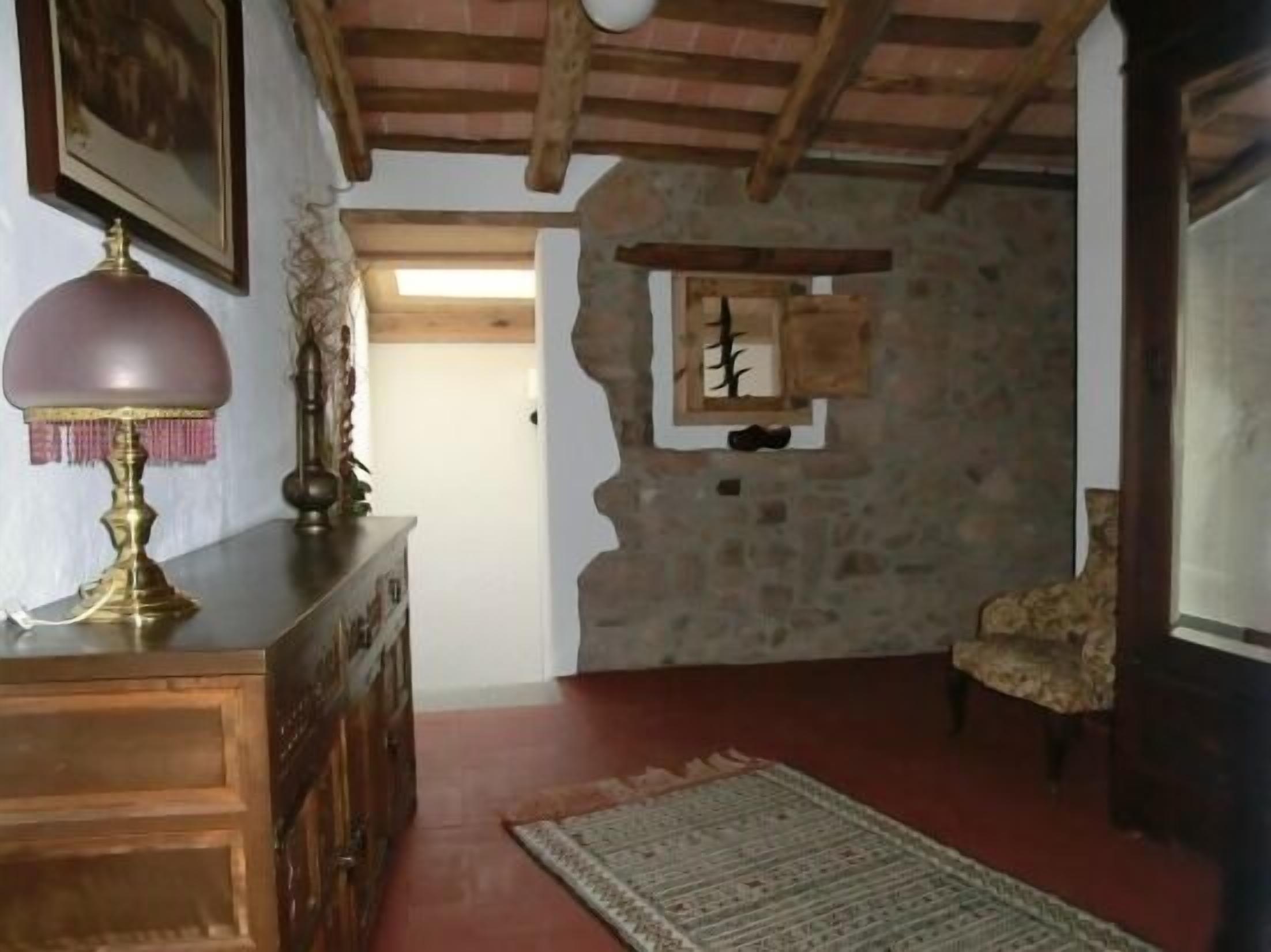 Una habitación rústica con paredes de piedra vista, techo de madera, un aparador de madera con una lámpara, una alfombra estampada y una silla.