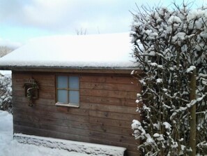 Una cabaña de madera con ventana y puerta, rodeada de nieve y árboles.