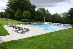 Piscina rectangular con solárium empedrado, rodeada de césped y árboles.