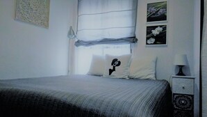 A bedroom with a bed, a nightstand with a lamp, a window with curtains, and a wall with a decorative piece and two framed pictures.