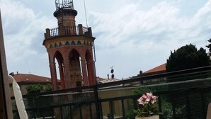 Un balcó amb dues cadires blanques i una taula petita, una planta en test i una vista d'una torre històrica amb arcs.