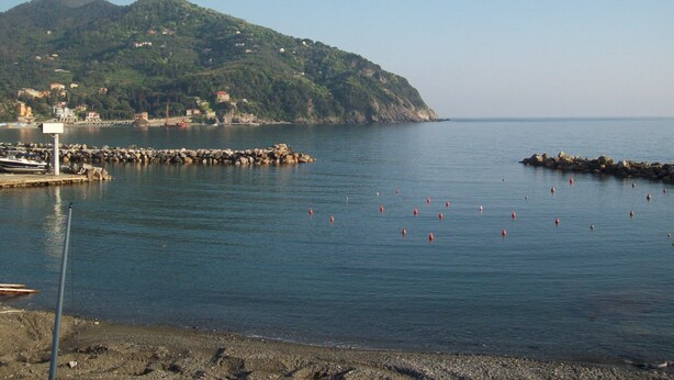 Escena costanera amb un bascul protector rocos, mar tranquil·la i un turó amb edificis.