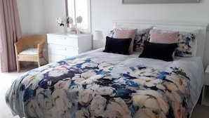 A bedroom with a floral bedspread, white furniture, a wicker chair, and a window with curtains. - Sea front Apartment In Castlerock, With Stunning Panoramic Views (Castlerock)