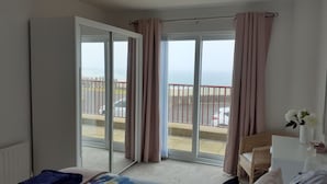 A bedroom with a bed, a small table with a vase, and a view of the sea through large sliding doors. - Sea front Apartment In Castlerock, With Stunning Panoramic Views (Castlerock)