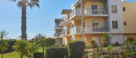 A multi-story residential building with balconies, surrounded by well-maintained gardens and palm trees.