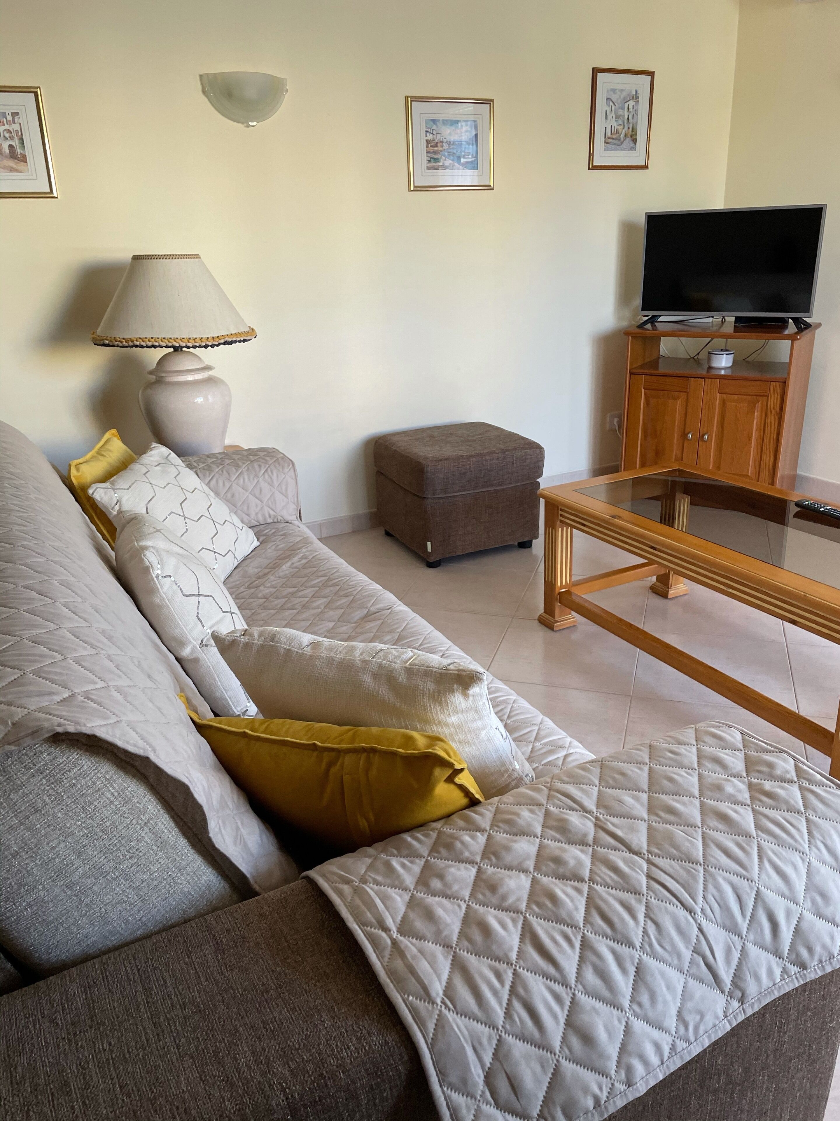 A living room with a sofa, ottoman, table, lamp, TV, and framed pictures on the wall.