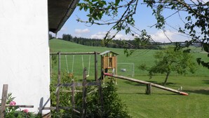 Una casita de juegos rústica de madera con columpios y un tobogán, rodeada de un campo verde exuberante y un edificio blanco.