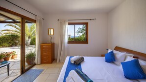 A bedroom with a large bed, a wooden headboard, a window with a view of greenery, and a door leading to an outdoor area with plants.