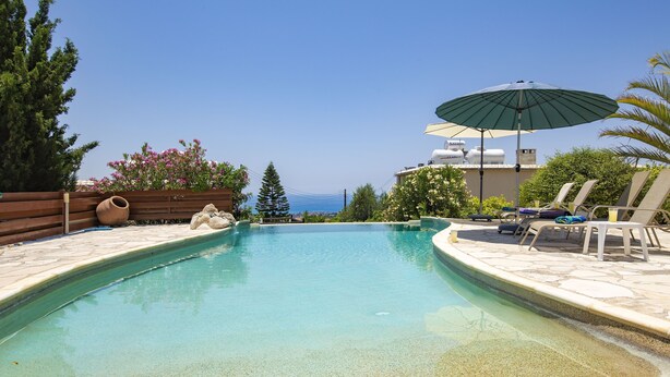 A swimming pool with lounge chairs and an umbrella, surrounded by greenery and a view of the sea.