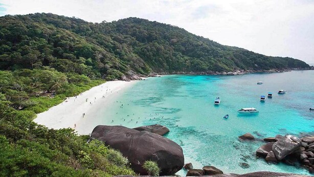 Una playa con aguas turquesas cristalinas, barcos y una gran roca al frente.