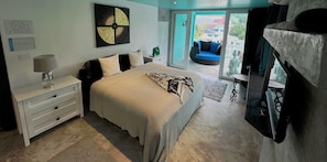 Quarto com cama, mesa de cabeceira, luminária, cômoda, televisão e vista para o exterior.