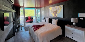 Quarto com uma cama grande, uma mesa de cabeceira, uma cadeira vermelha e vista para uma área externa através de uma porta de vidro.